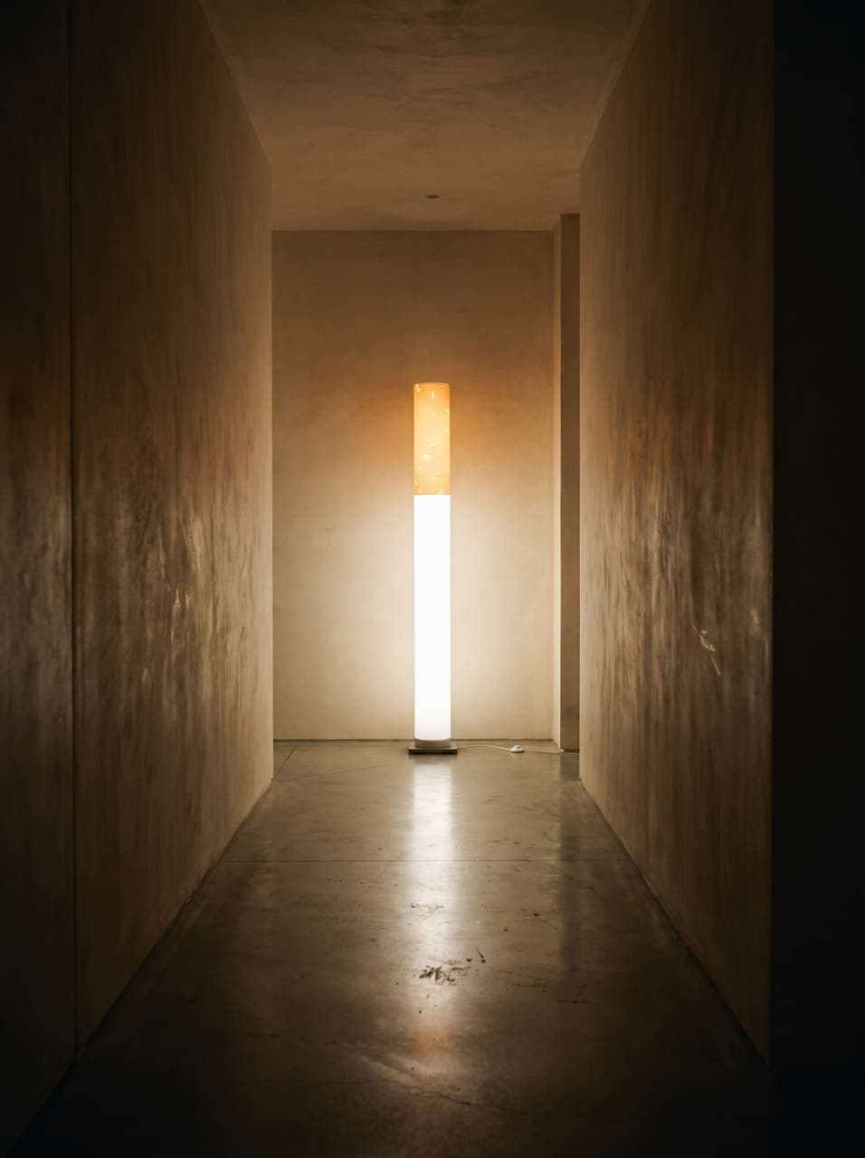 A long limewashed corridor with the lit floor lamp at its far end, the rest in soft shadow