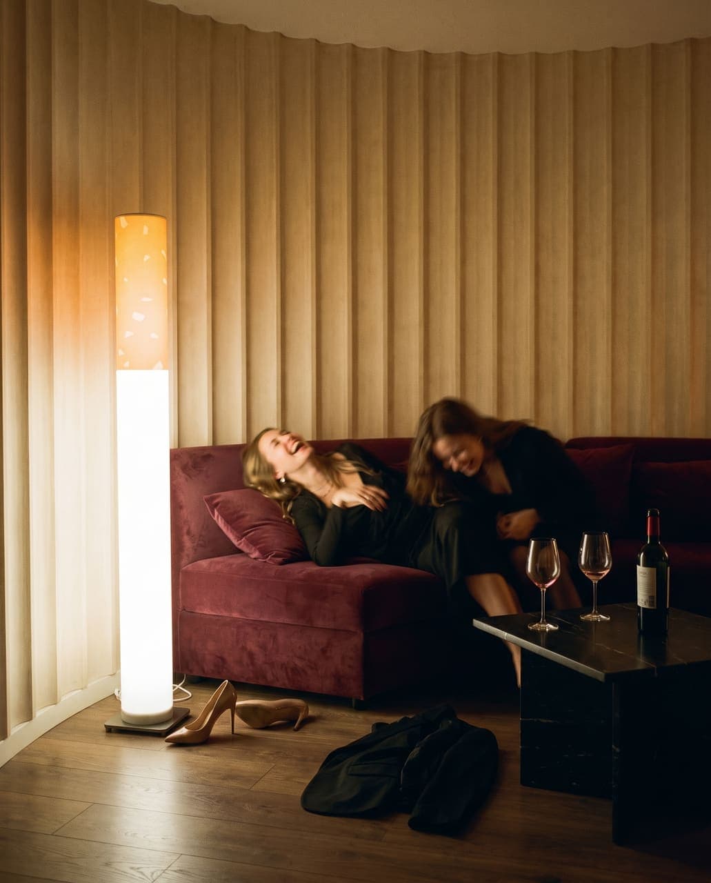 Two friends collapsed laughing on a burgundy velvet sofa with heels kicked off and glasses on the table, the floor lamp beside them