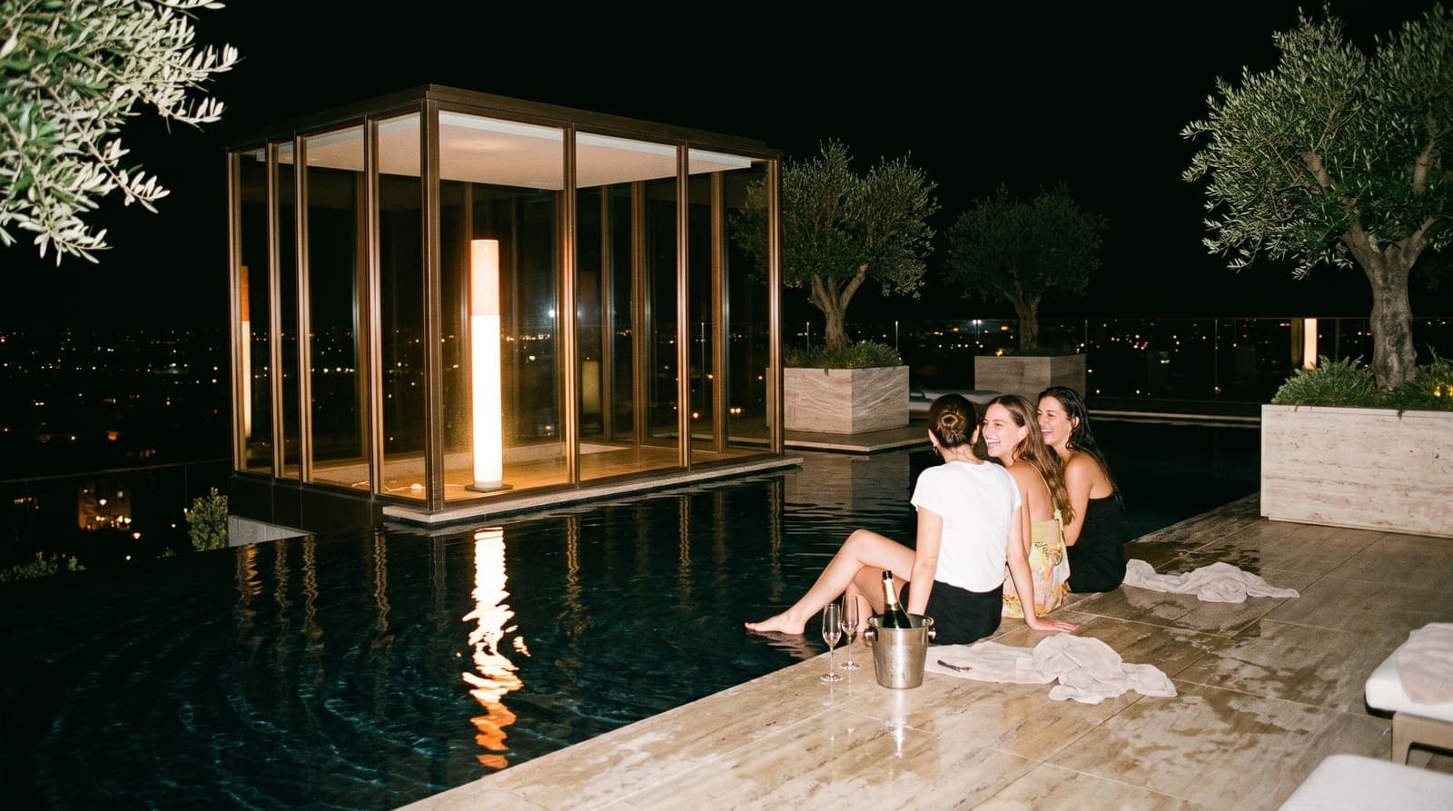 Three friends on the edge of a rooftop pool at night with their legs in the water, the floor lamp glowing inside the glass pavilion behind