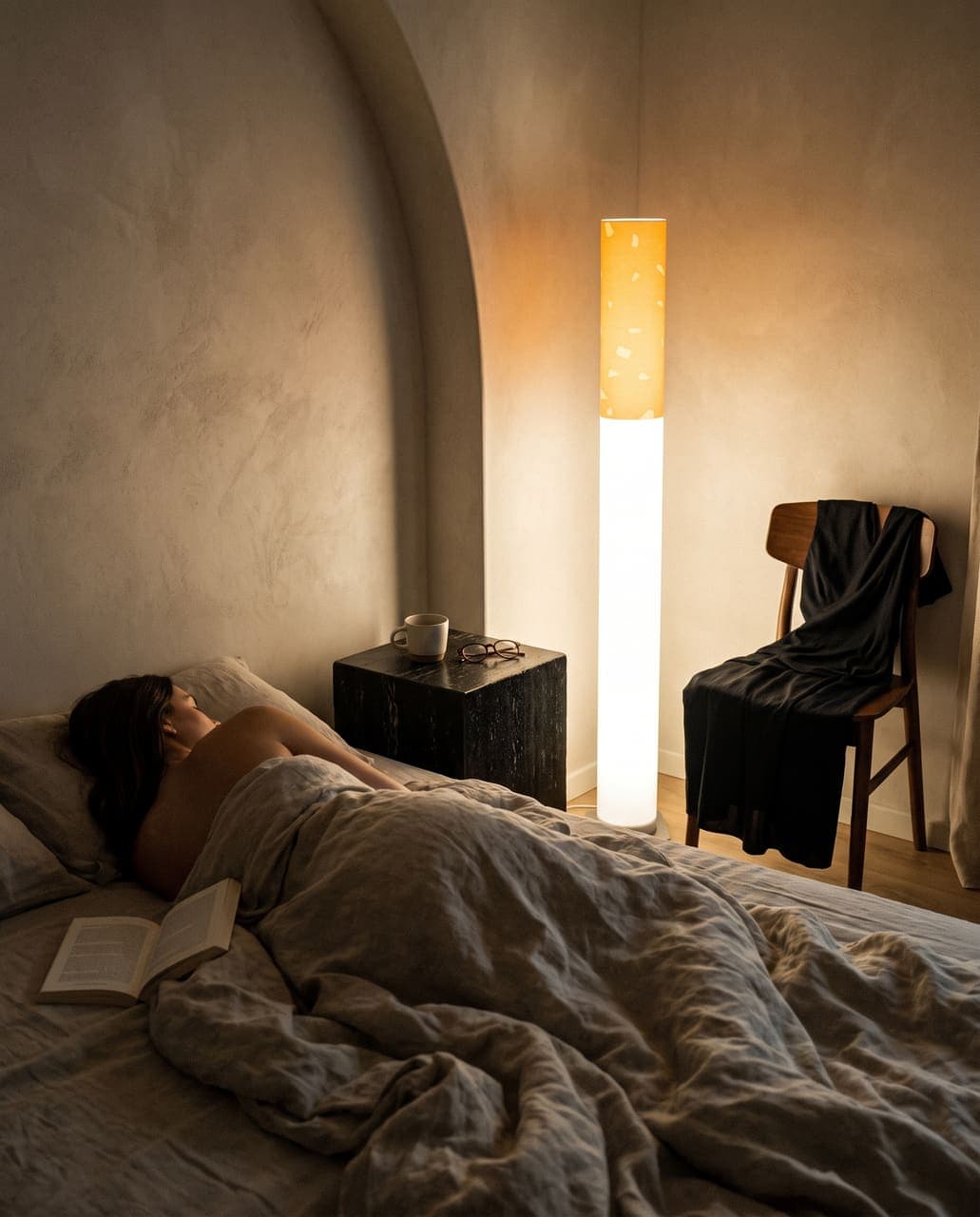 A woman asleep face down in rumpled linen with a book fallen open beside the bed, the floor lamp still on in the corner