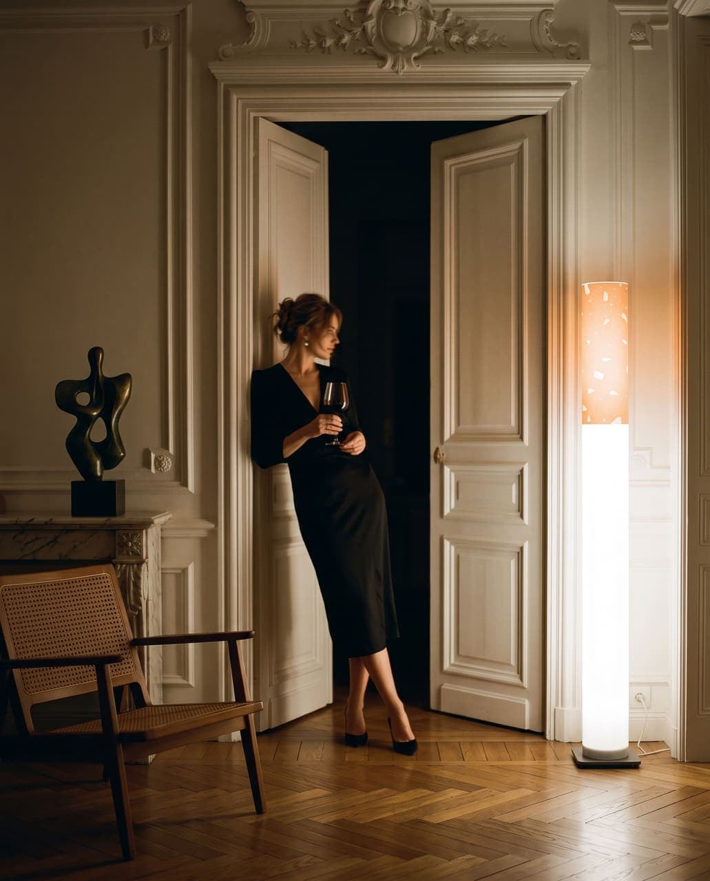 A woman in a black dress leaning in a tall doorway of chalk-white mouldings with a glass of wine, the floor lamp glowing beside her