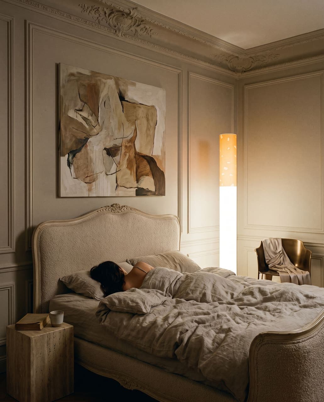 A woman asleep in an antique bed reupholstered in boucle under tall painted mouldings, the floor lamp still on in the corner
