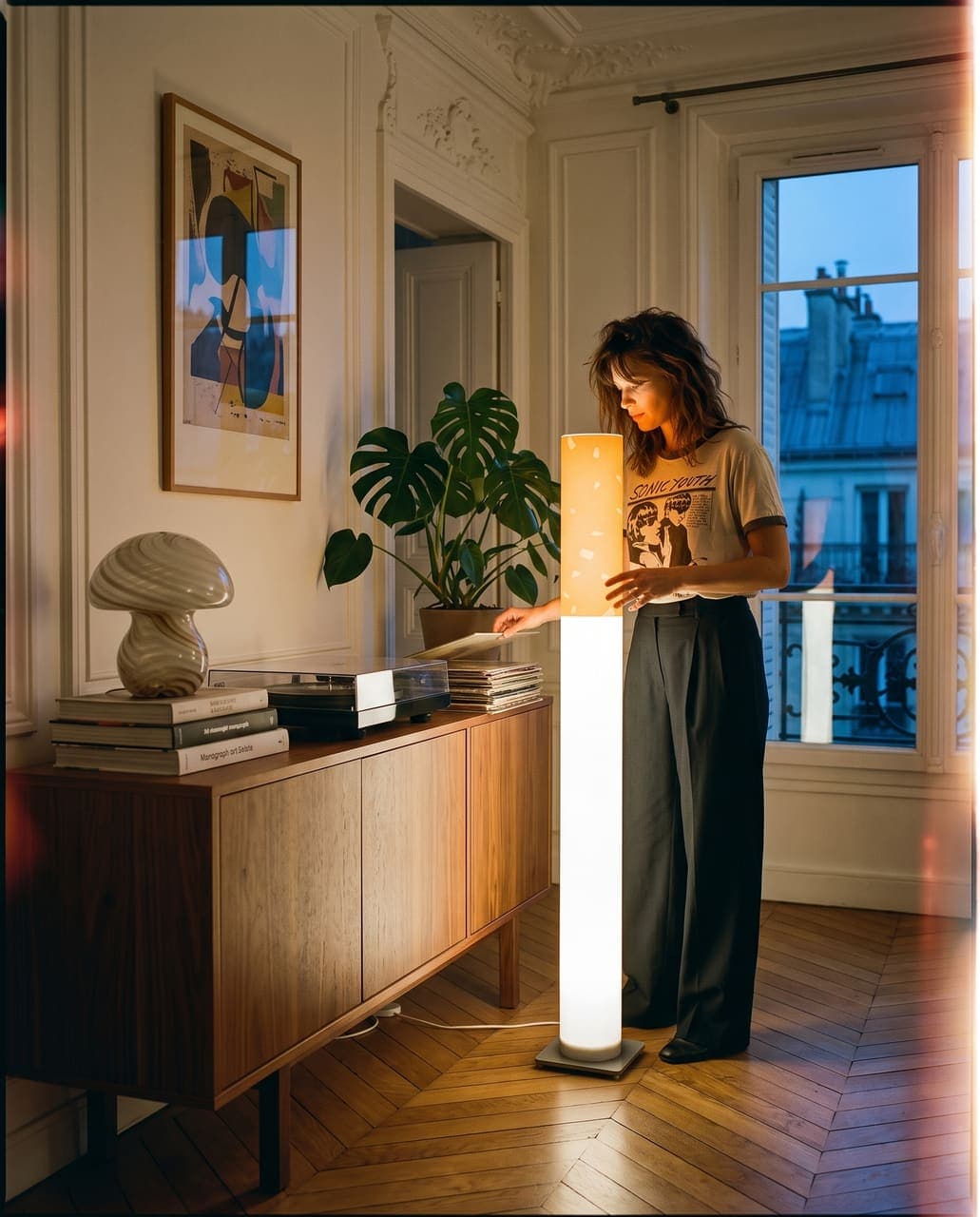 A woman in a band T-shirt flipping through records at a walnut sideboard beneath a monstera, the floor lamp glowing beside her