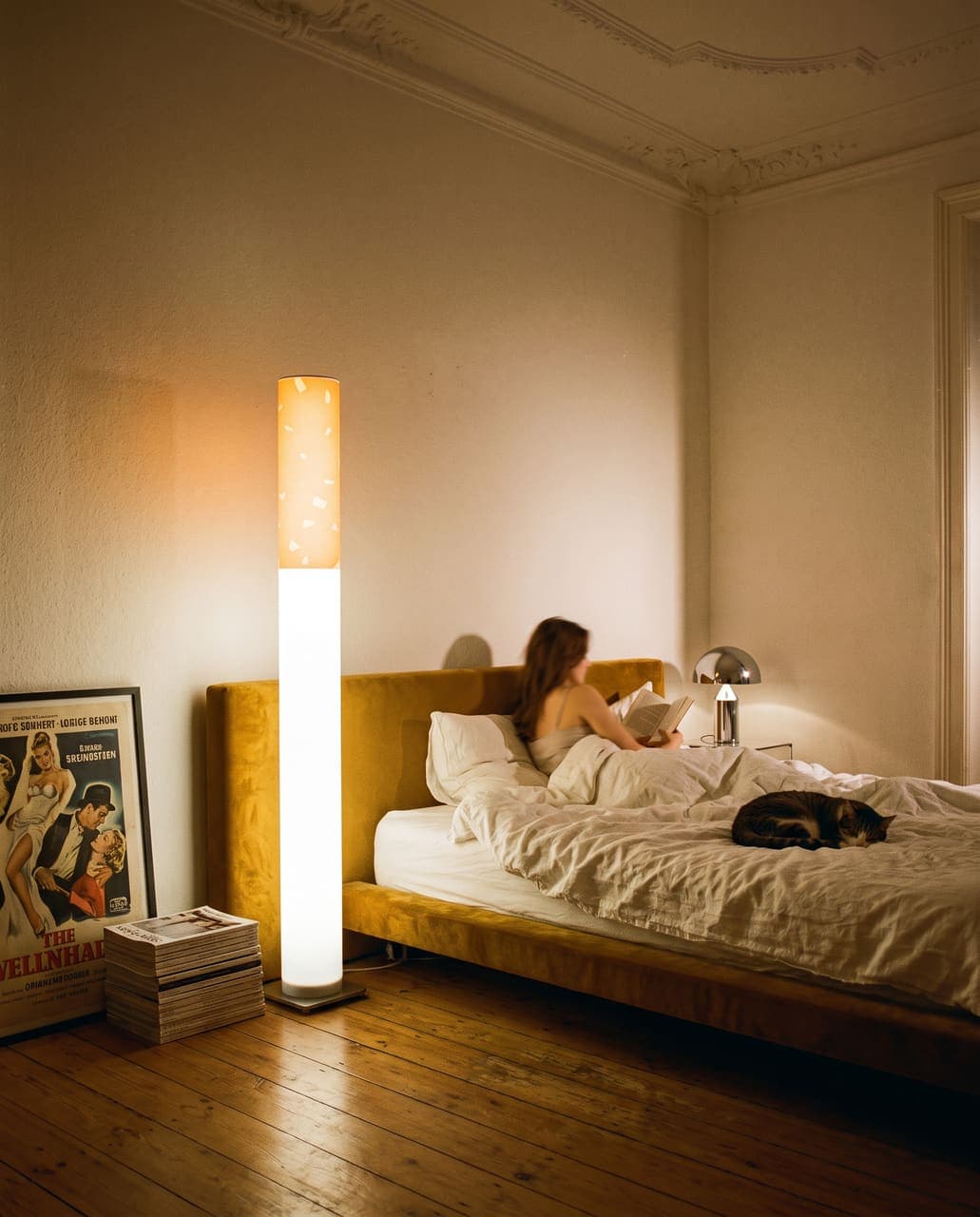 A woman reading in bed against a mustard velvet headboard with a cat on the duvet and magazines on the floor, the floor lamp on in the corner