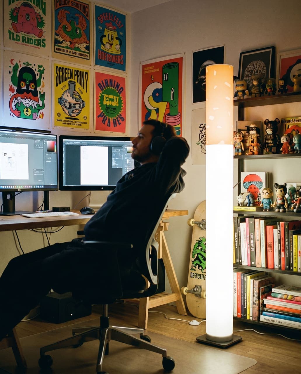 A designer stretching back in his chair under a wall of screen-printed posters with two monitors glowing, the floor lamp beside the desk