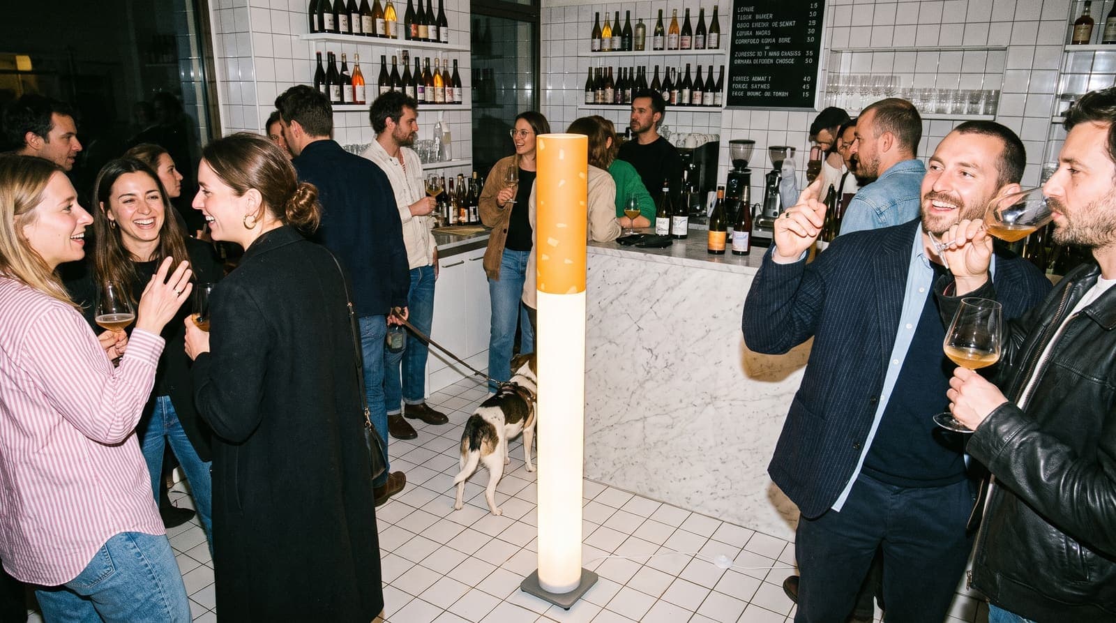 A crowded natural wine bar with white tiles and a chalkboard menu, a dog on a lead, the floor lamp standing on the floor by the counter