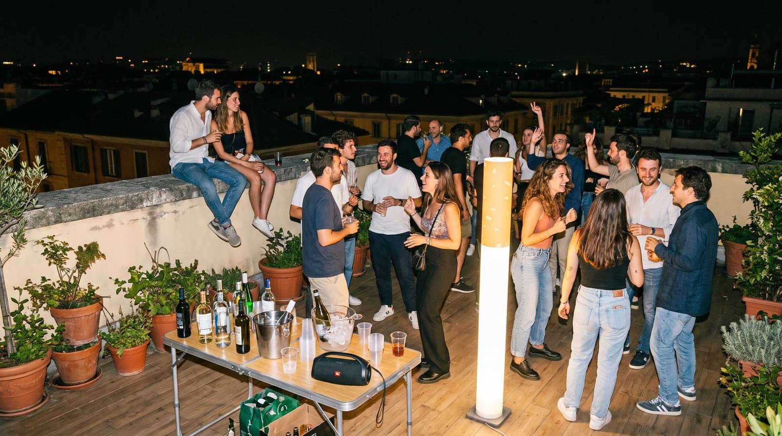 Friends at a rooftop party at night around a folding table with bottles in an ice bucket, the floor lamp glowing among them
