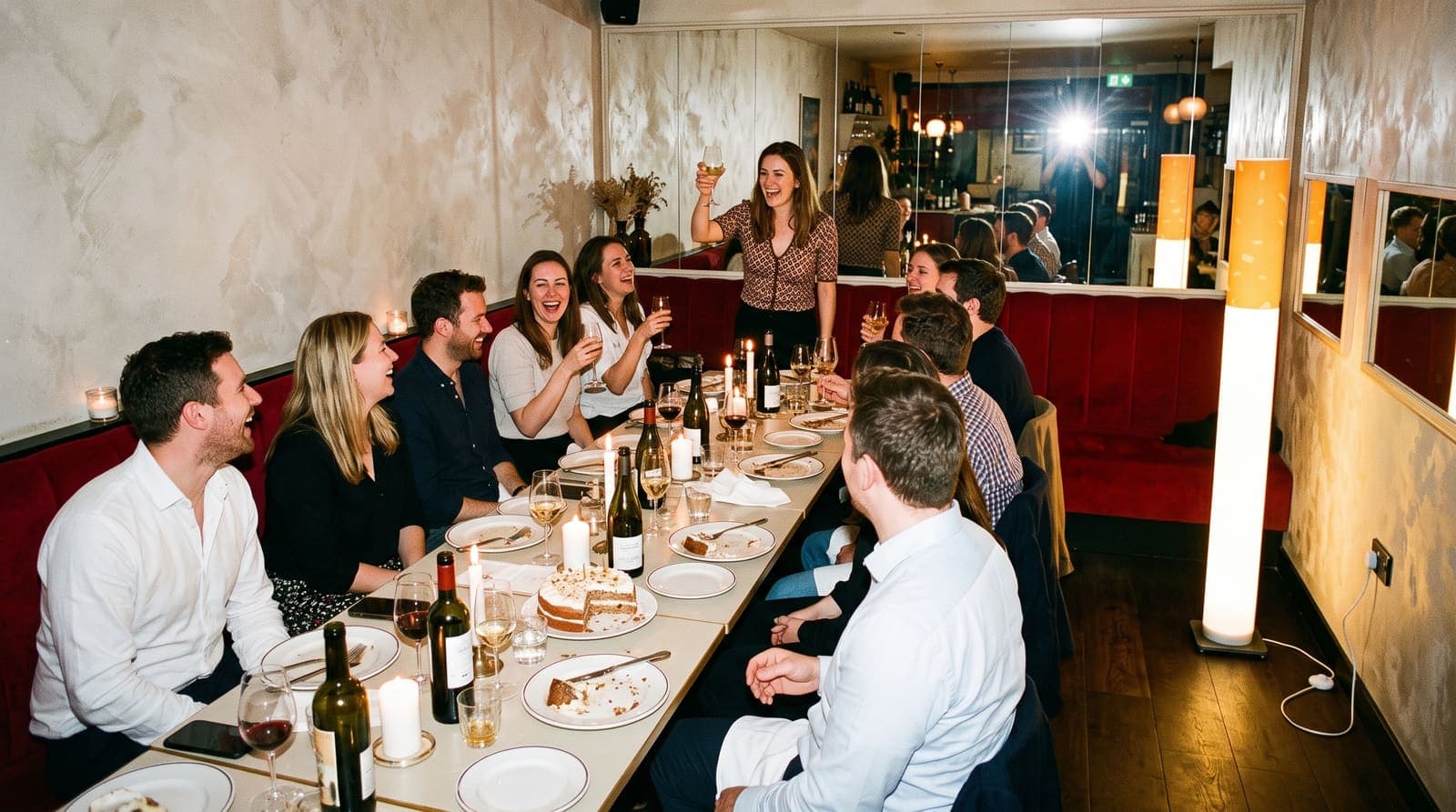 Twelve friends raising glasses around a long table with a half-eaten cake and burnt-down candles, the floor lamp glowing in the corner