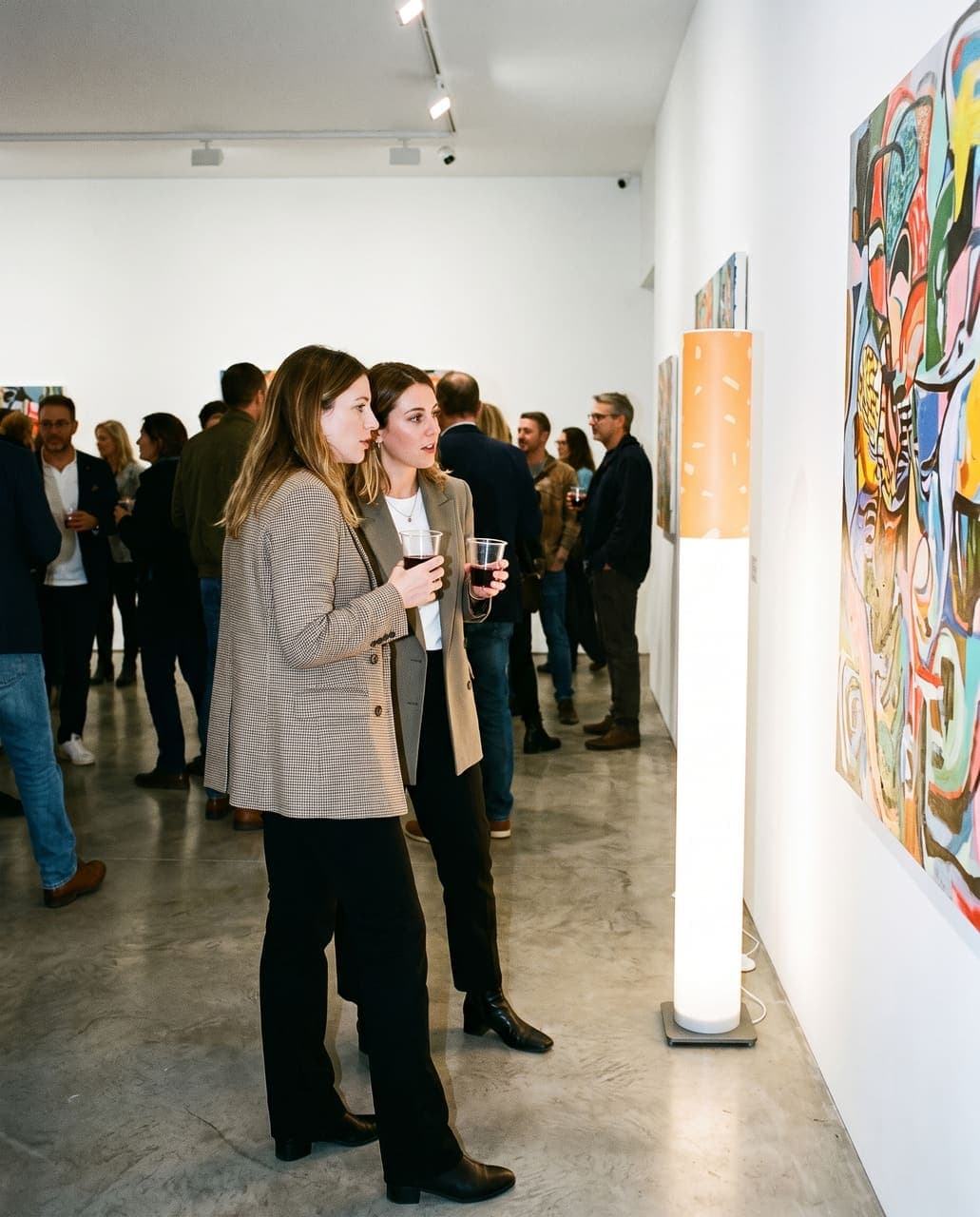 Two women talking in front of a large painting at a gallery opening, the floor lamp glowing beside them