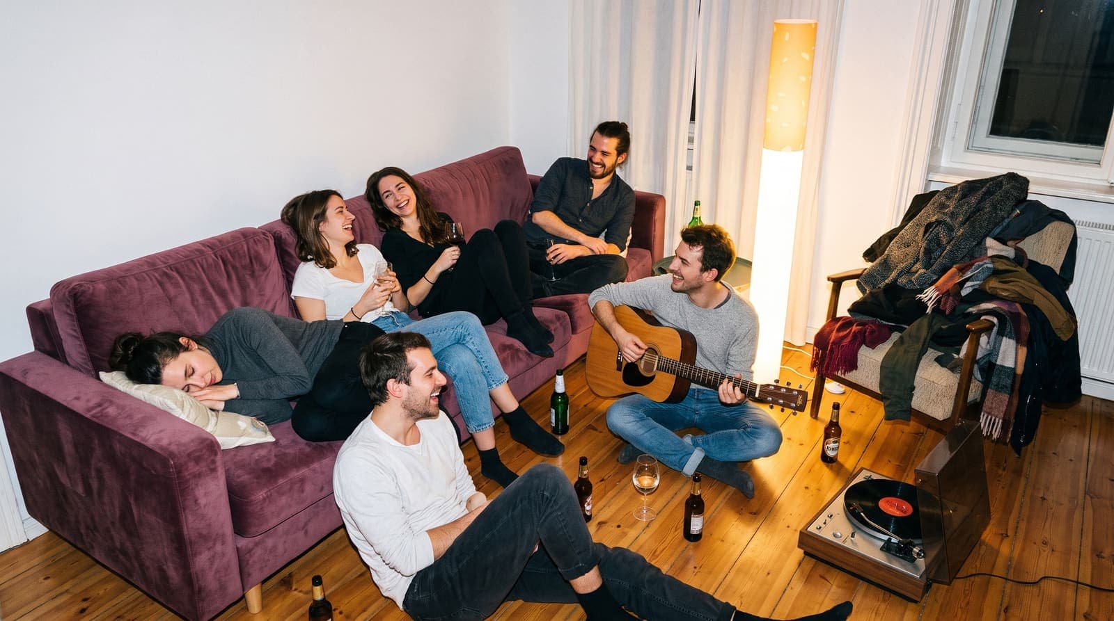 An afterparty at three in the morning with someone playing guitar on the floor and someone asleep on the sofa, the floor lamp still on