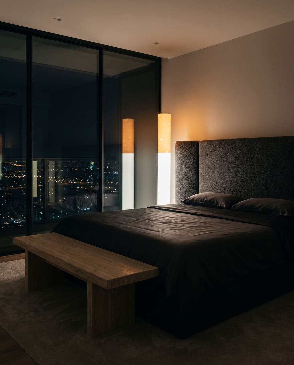 The lit floor lamp in a dark bedroom with a boucle headboard, silk bedding and a city view