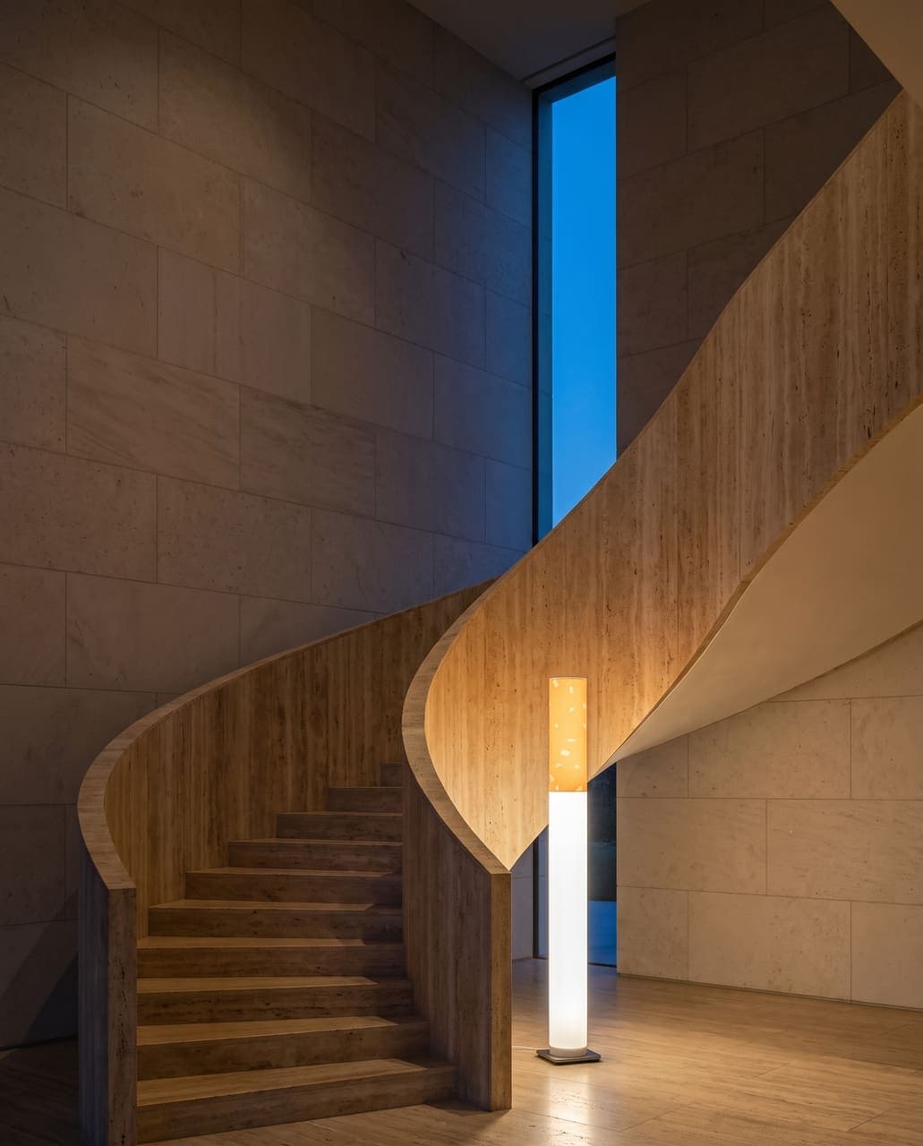 The floor lamp glowing at the foot of a sweeping travertine staircase in a limestone entrance hall at dusk
