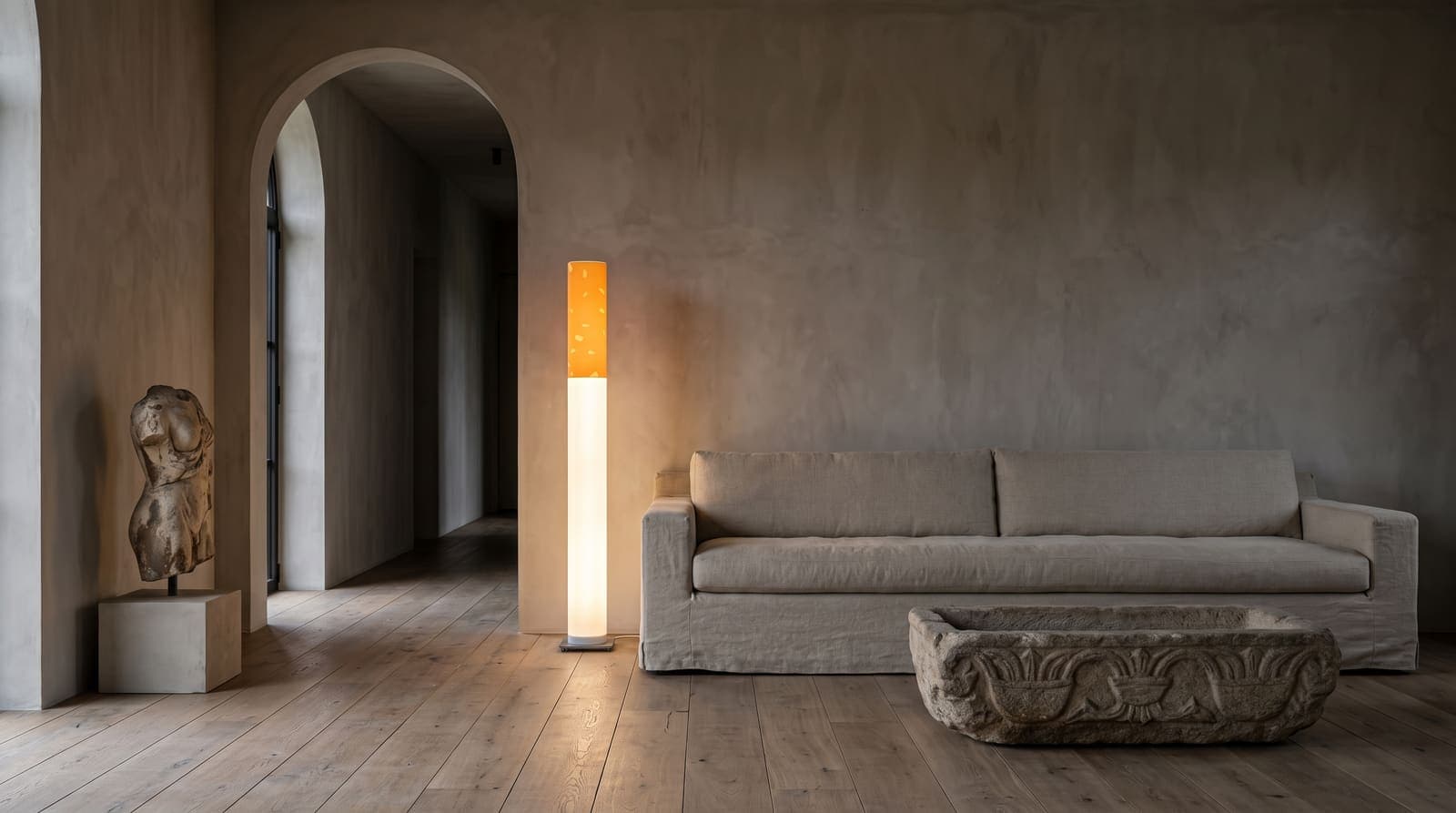 The lit floor lamp beside a long linen sofa in a vast room of raw plaster, reclaimed oak and an antique stone trough table