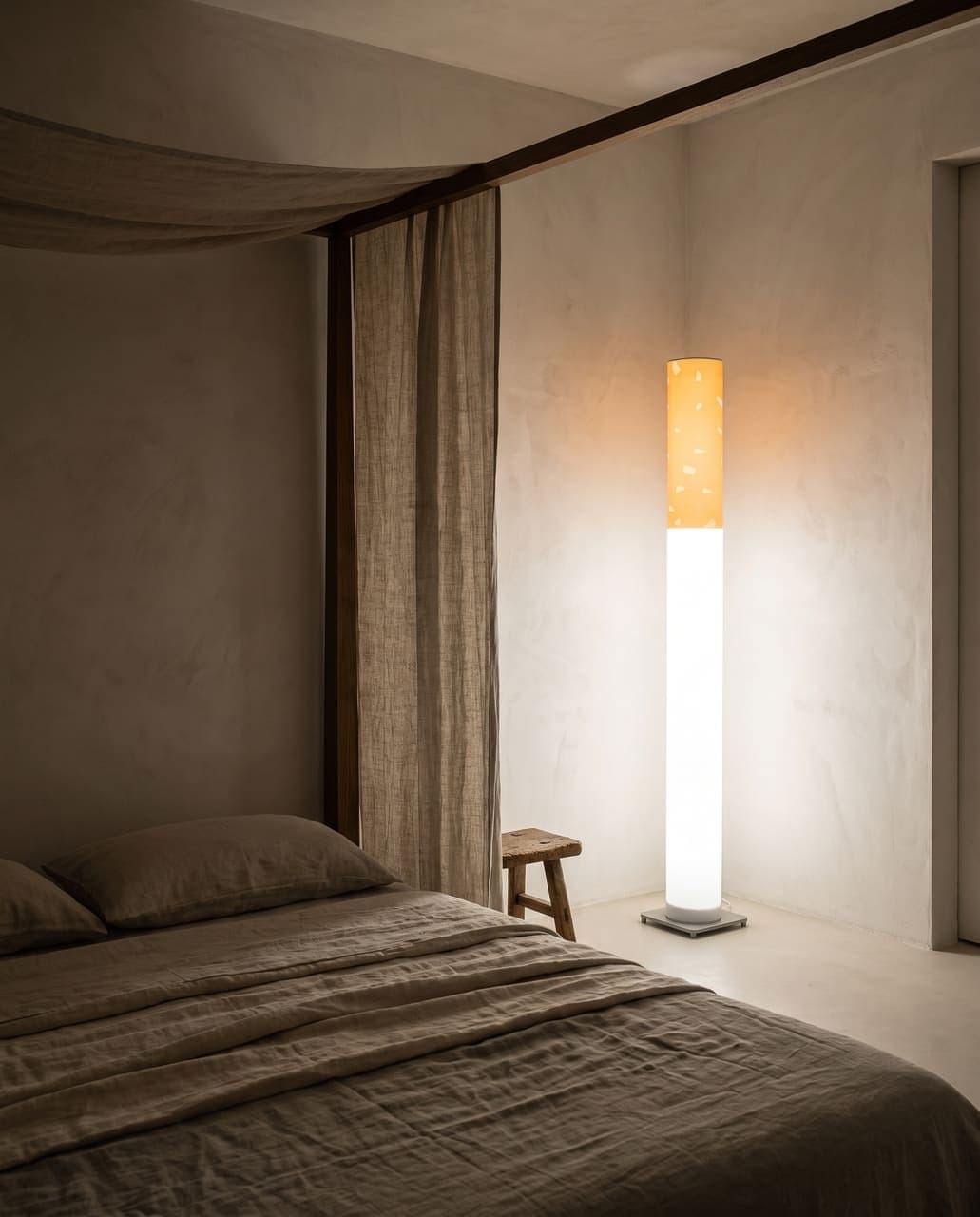 The floor lamp glowing in a bedroom of raw plaster walls, a dark oak canopy frame and layers of heavy linen