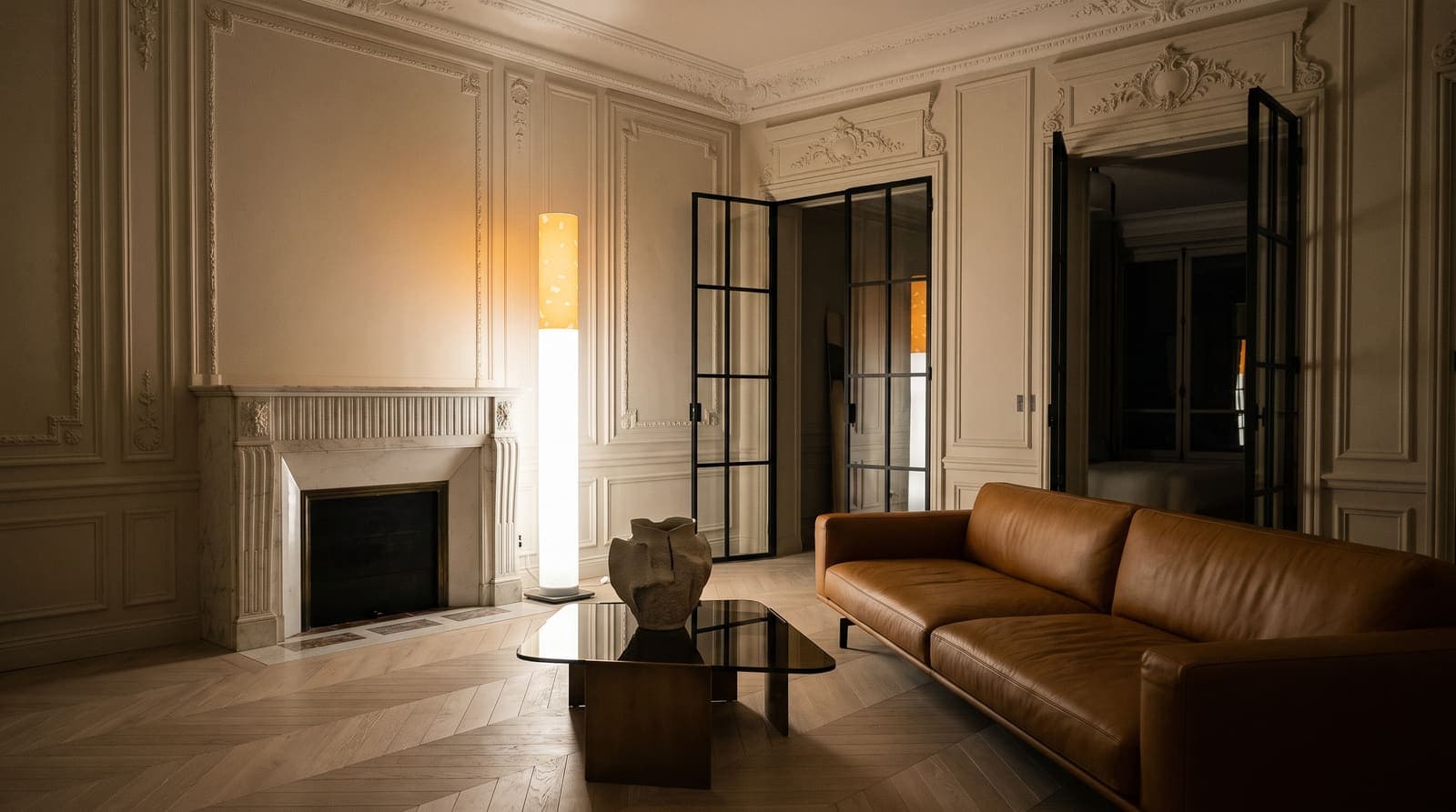 The lit floor lamp by a fluted marble fireplace in a Paris apartment with pale stone mouldings, black steel doors and a caramel leather sofa