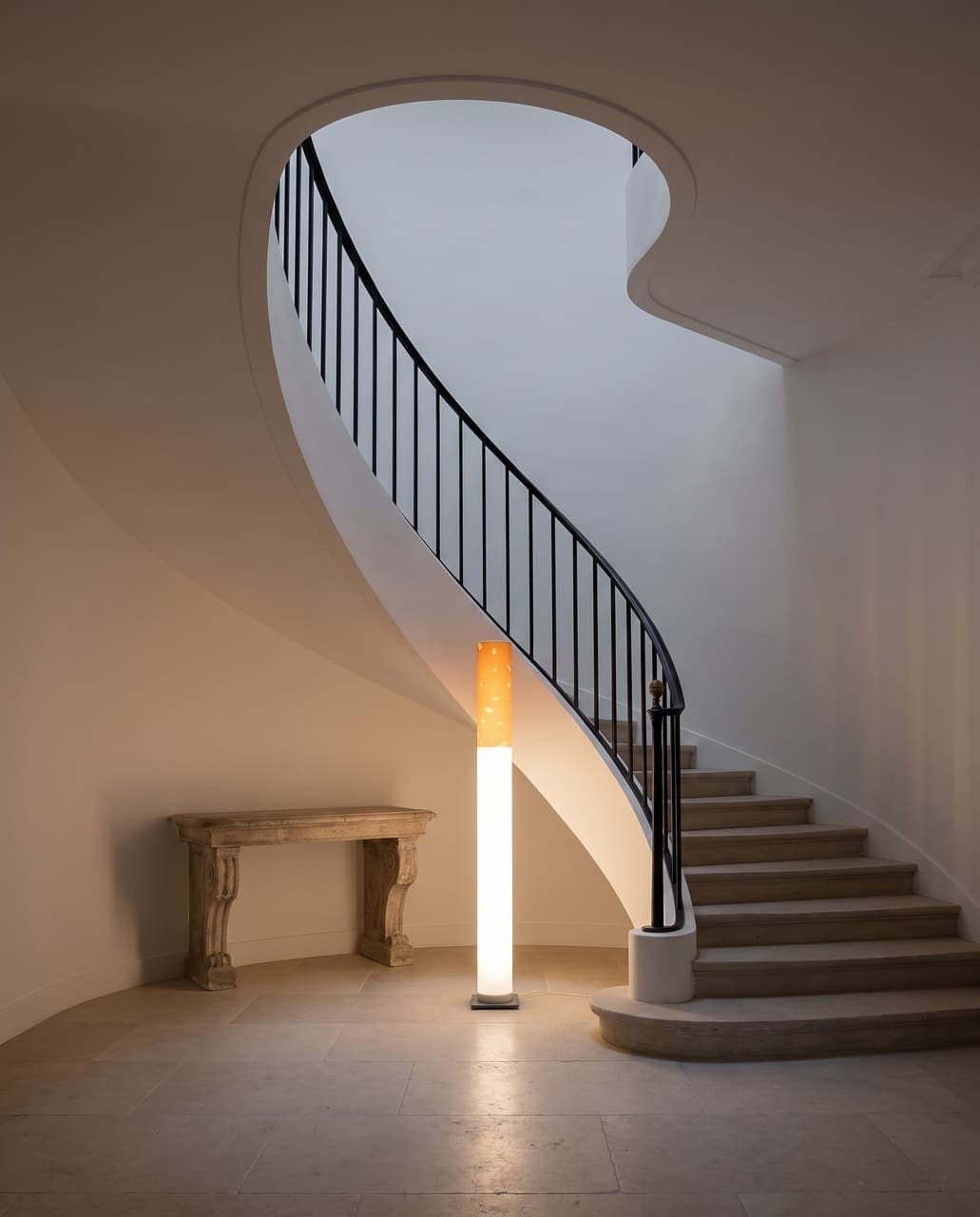 The floor lamp glowing at the foot of a curved staircase with a black steel balustrade and white plaster walls
