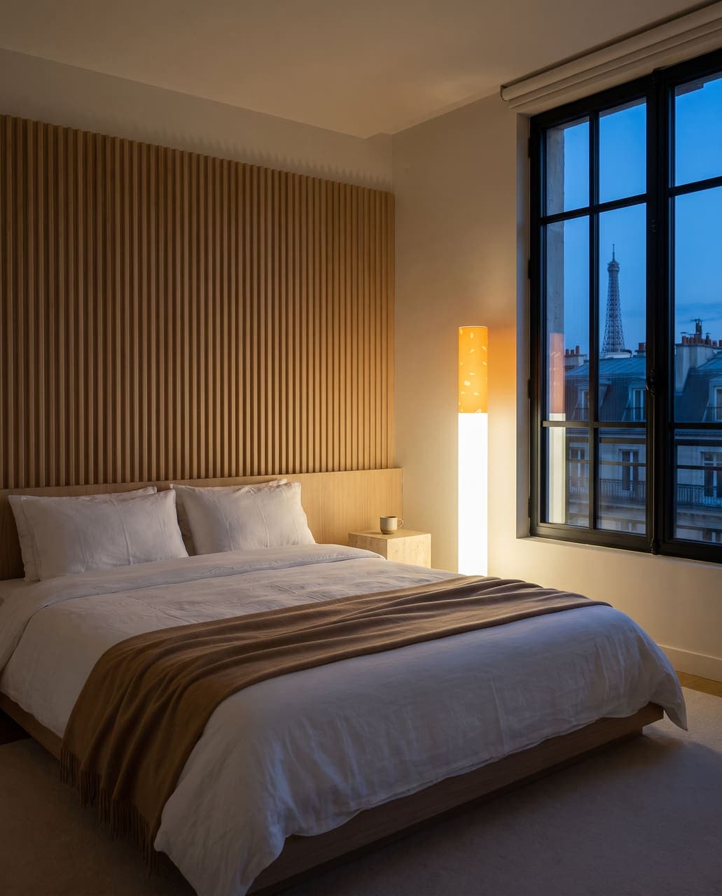 The floor lamp glowing in a bedroom with a fluted oak headboard wall, white linen and Paris rooftops in the window