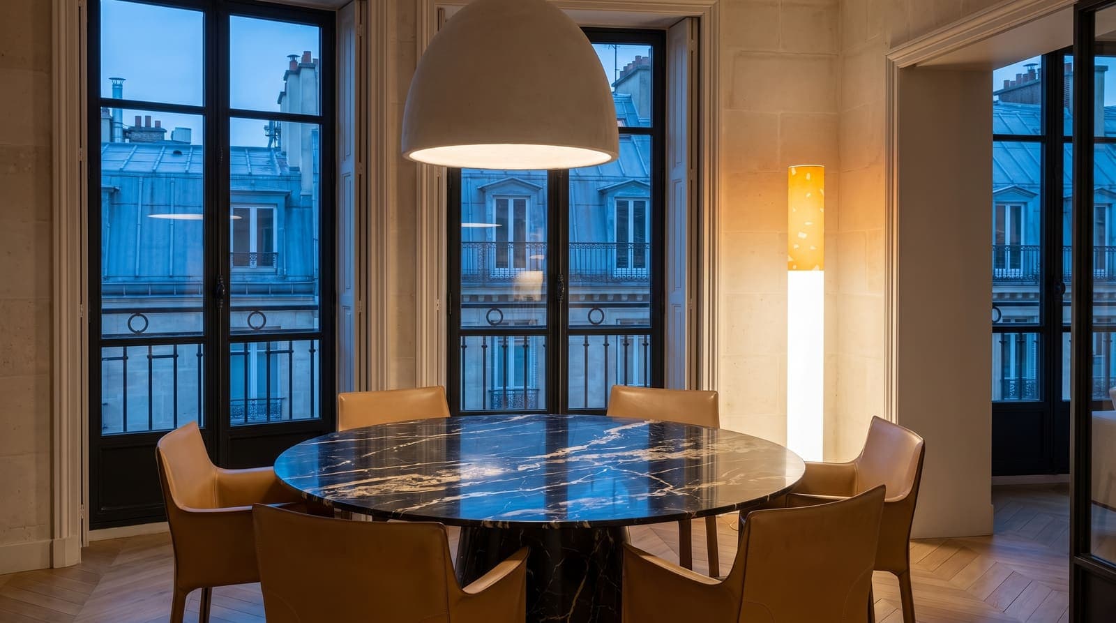 The lit floor lamp beside a round black marble dining table and tan leather chairs, zinc rooftops at dusk through tall windows