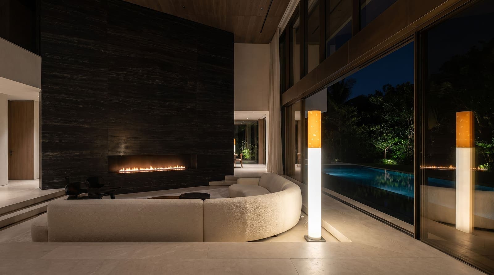 The floor lamp glowing at the edge of a sunken cream boucle lounge in a double-height room with a black travertine wall and a lit pool outside