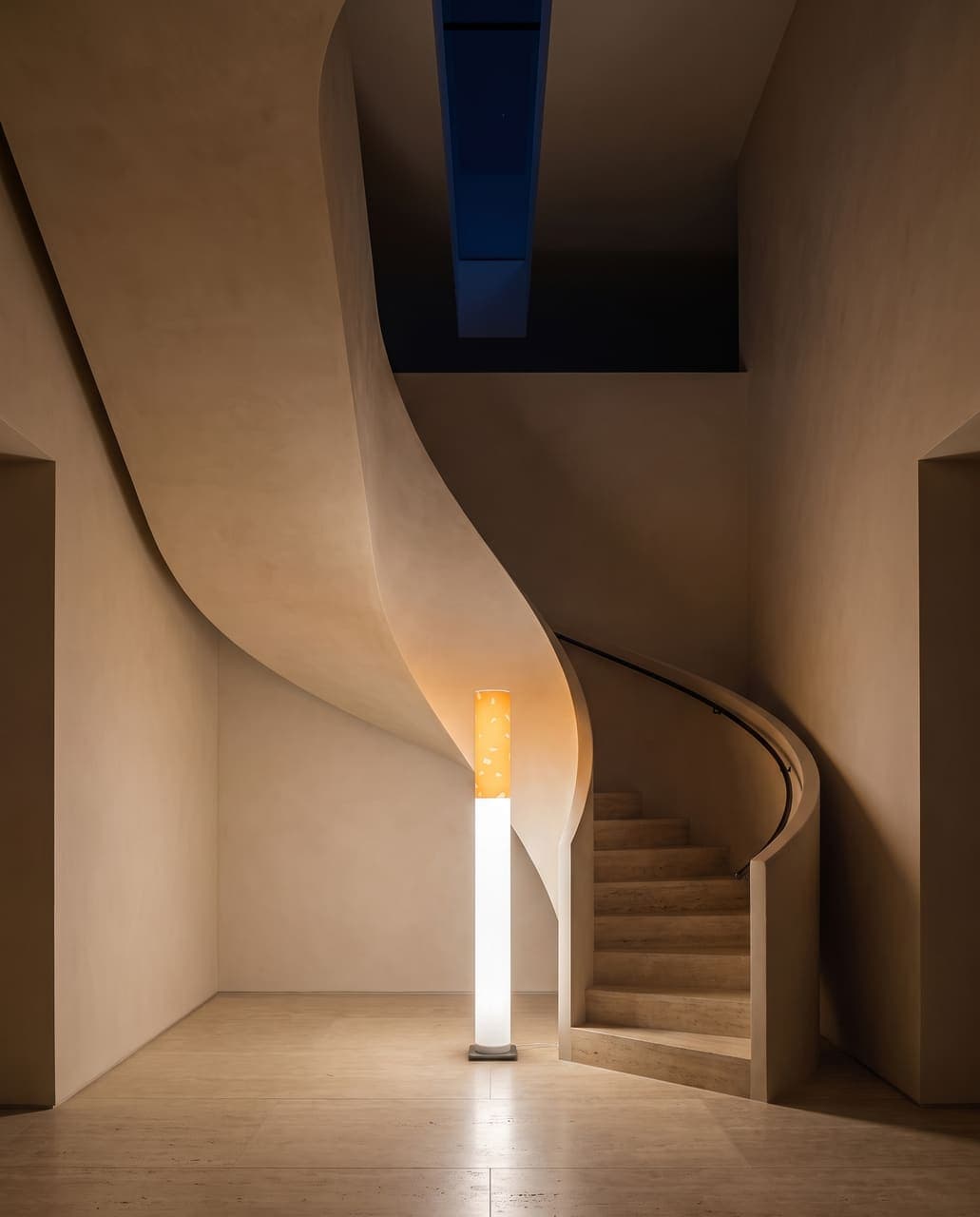 The lit floor lamp at the base of a seamless polished-plaster spiral staircase rising through a double-height void