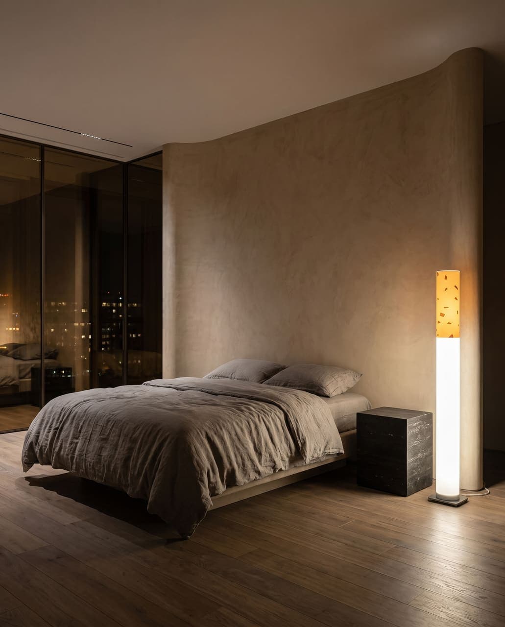 The floor lamp glowing beside a low floating bed against a curved plaster wall, bronze glass and city lights beyond