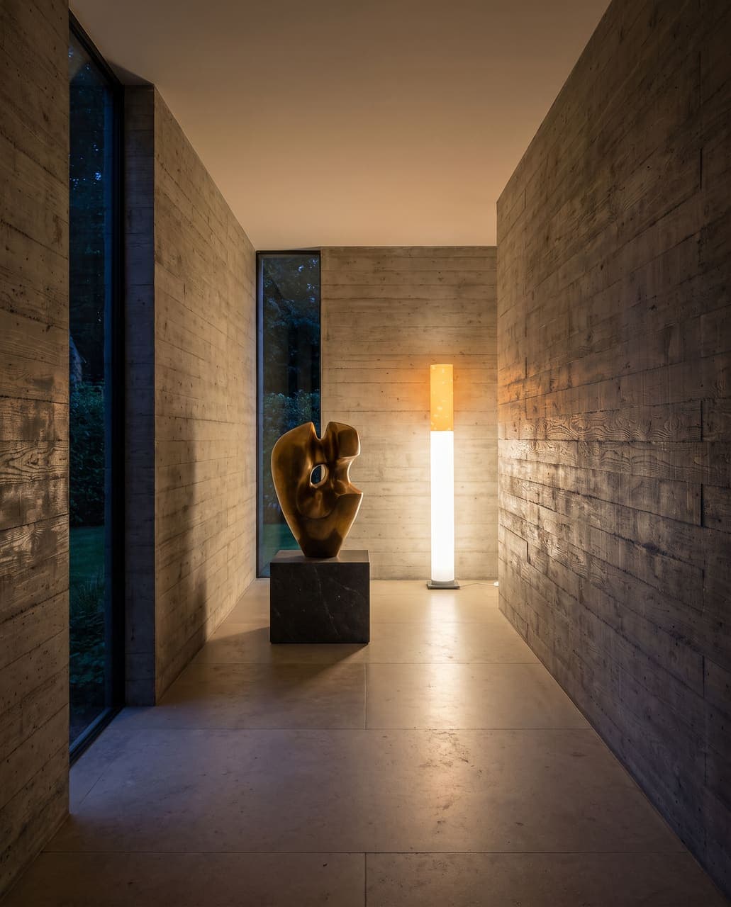 A board-formed concrete corridor with a bronze sculpture on a plinth and the lit floor lamp at the end