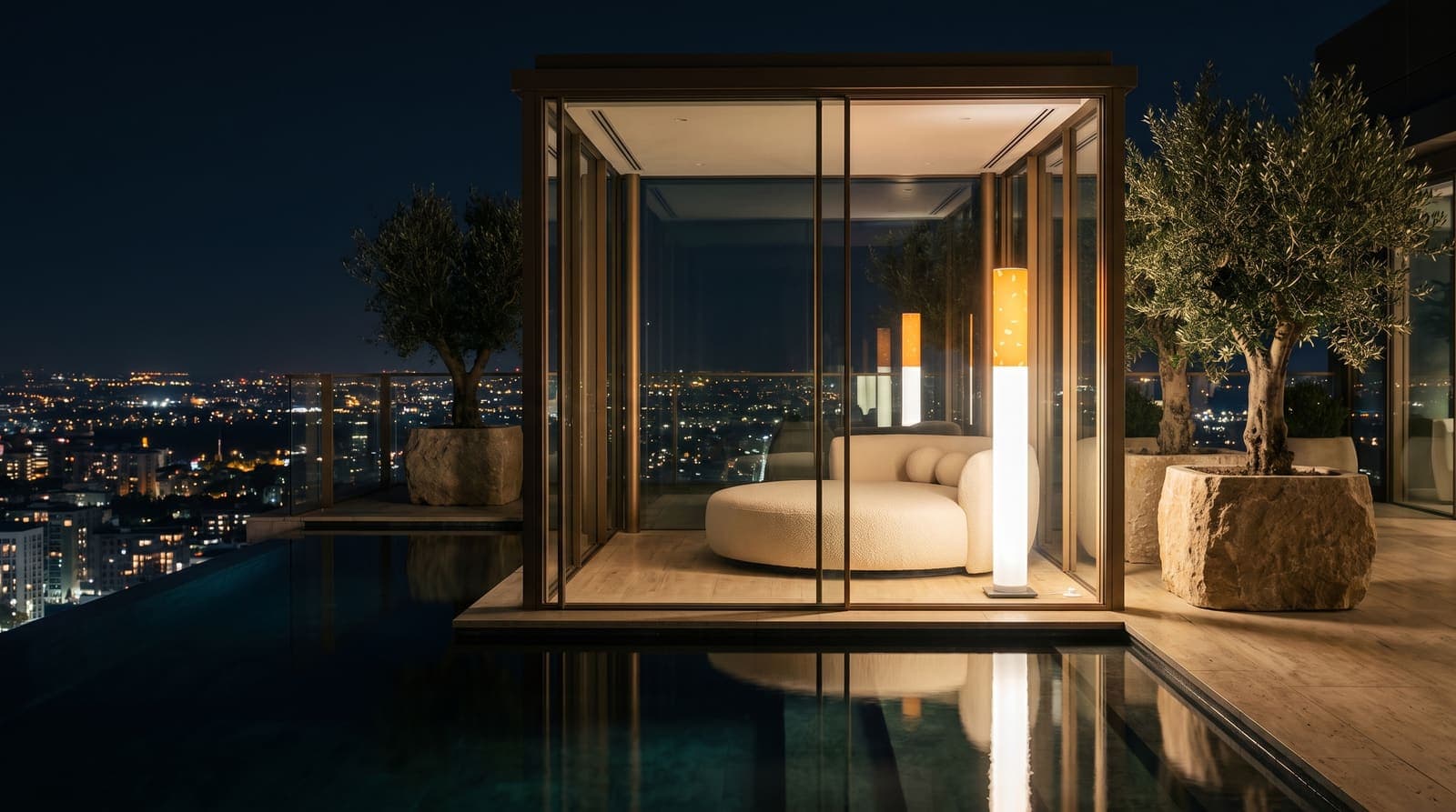 The floor lamp glowing inside a glass rooftop pavilion beside a curved daybed, a dark pool running inside to out and the city far below