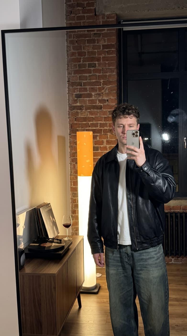 A man in a leather jacket taking a mirror selfie in a brick loft, the floor lamp glowing behind him beside a record player