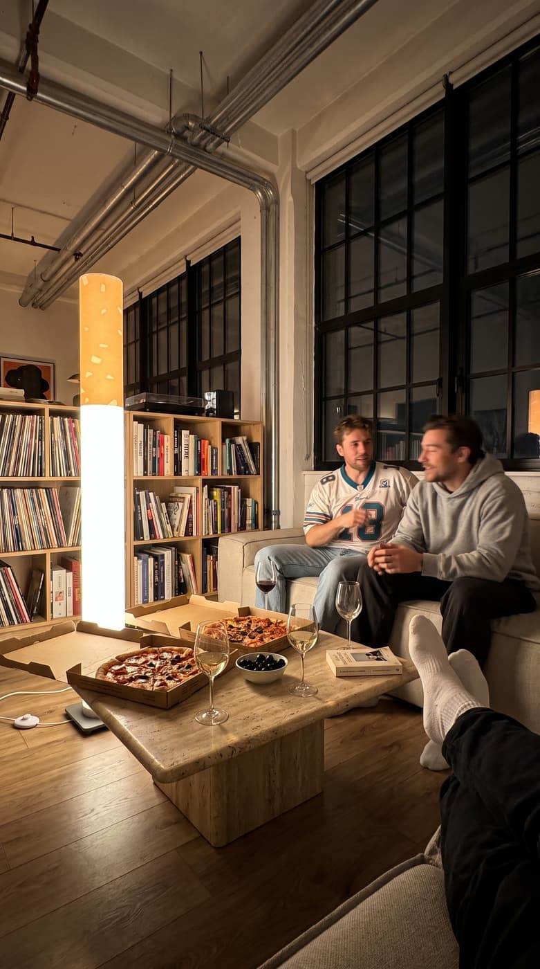 Two friends on a sofa with pizza boxes and wine on a travertine table in a loft, the floor lamp glowing beside the bookshelves