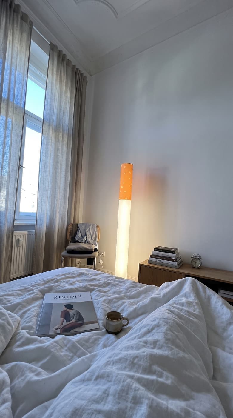A magazine and a cup on white linen in a sunlit bedroom, the floor lamp glowing softly by the curtain
