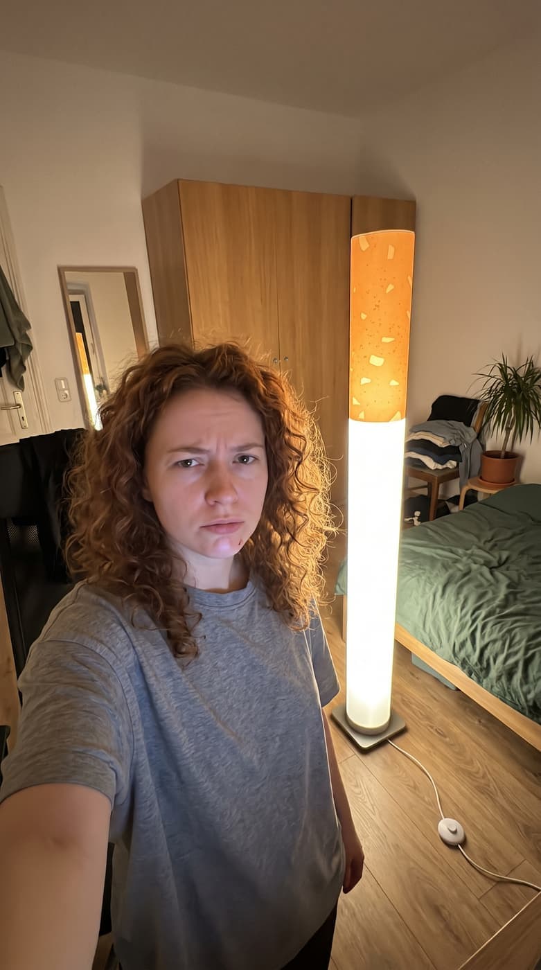 A woman with curly hair pulling a face in a selfie beside the glowing floor lamp in her bedroom