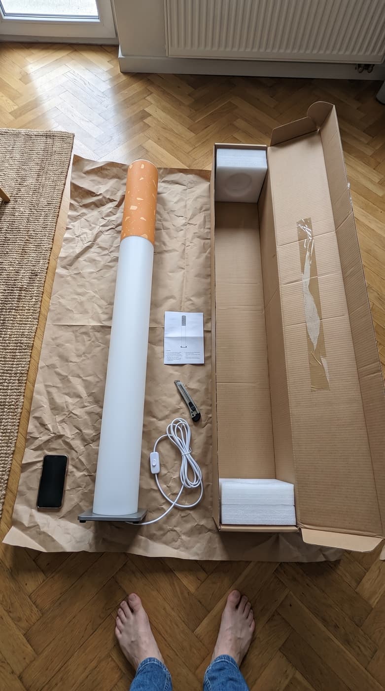 The floor lamp laid on brown packing paper beside its long cardboard box, the cord and the manual next to it, seen from above