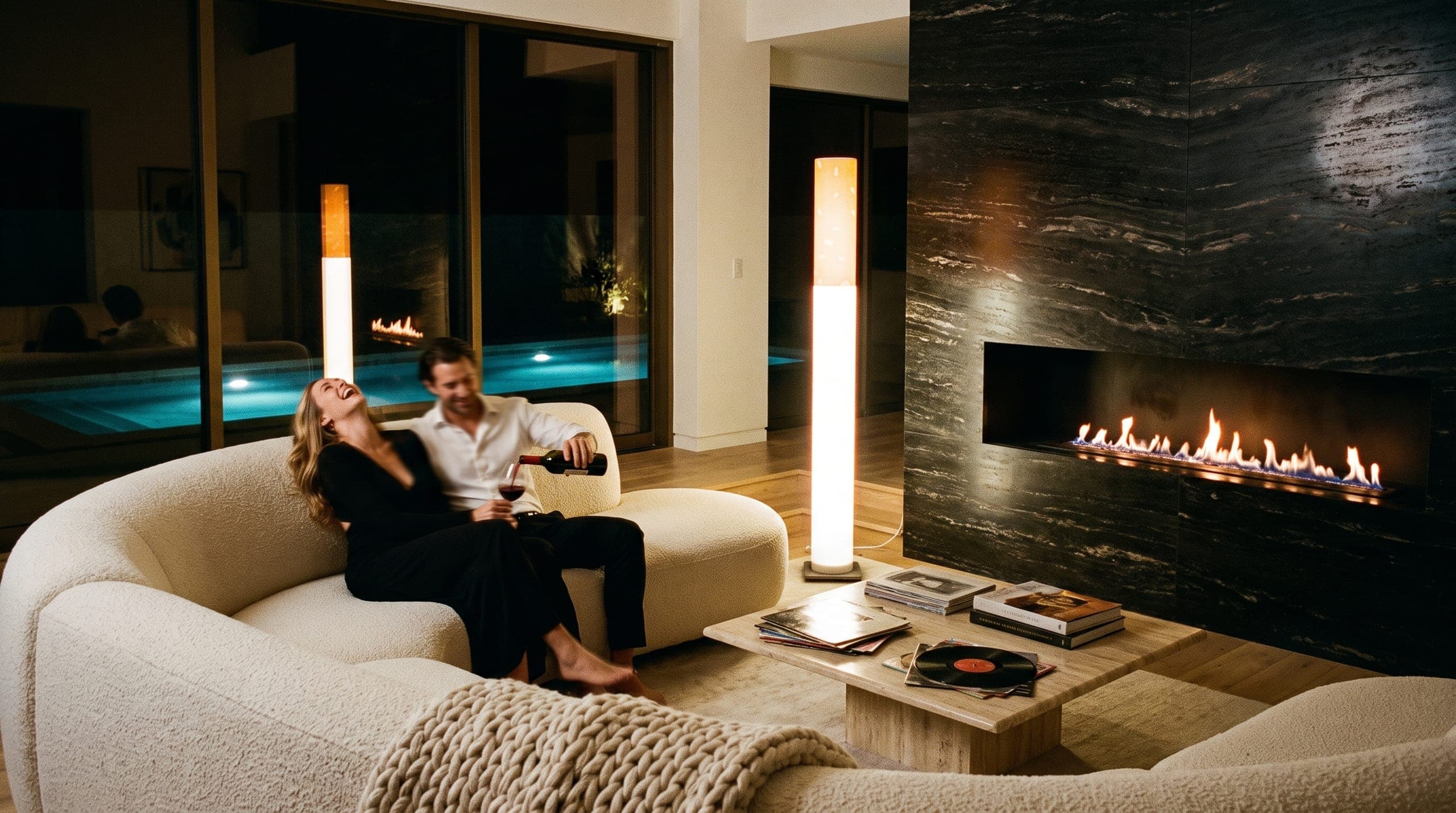 A couple on a curved cream sofa by a long fireplace slot at night, she laughing, he pouring wine, the floor lamp glowing at the edge of the sunken lounge